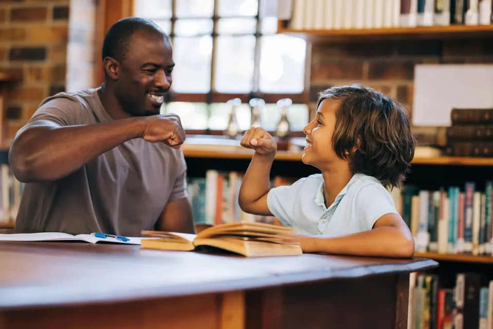 A student and an adult sharing a fist bump across a library table
