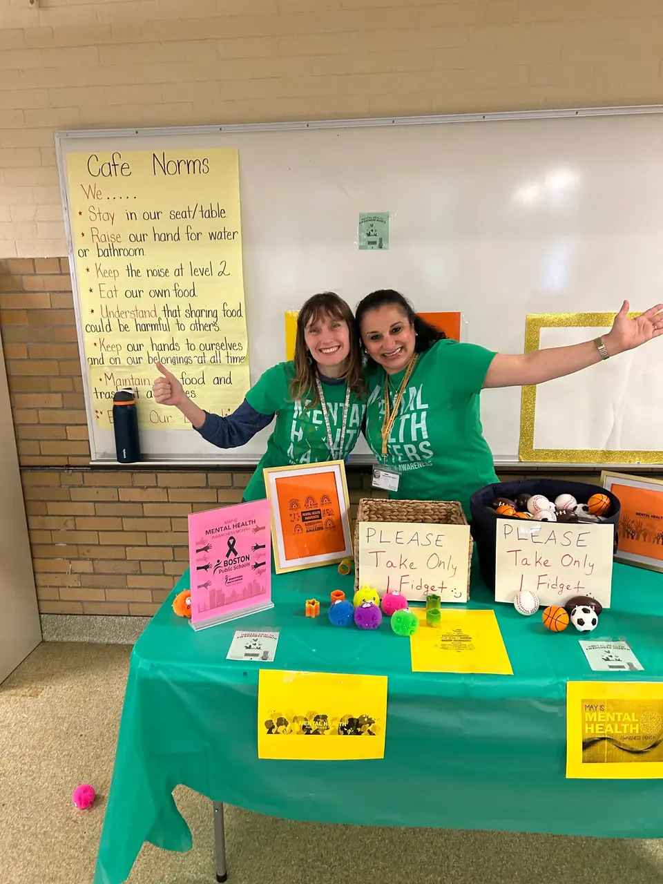 Two Behavioral Health Services staff members in green Mental Health Awareness Month shirts behind an activity table in a school cafeteria