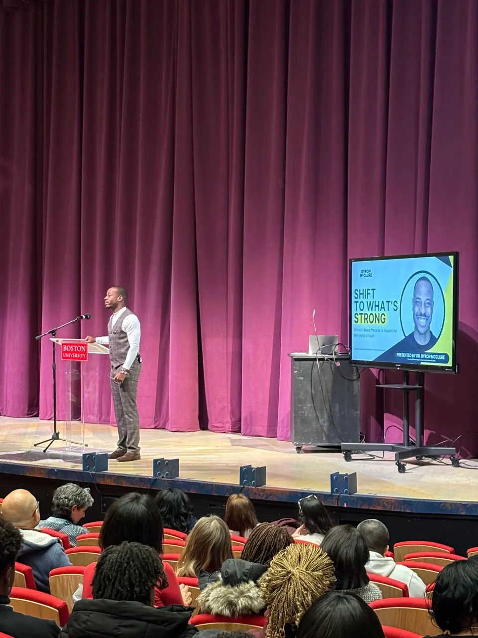 Dr. Byron McClure at a Boston University lectern on a theater stage, with his title slide Shift to What's Strong on a screen beside him and an audience in the foreground