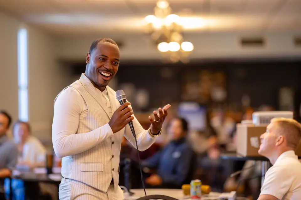 Dr. Byron McClure speaking into a handheld microphone and smiling while presenting to Boston school psychologists seated at round tables